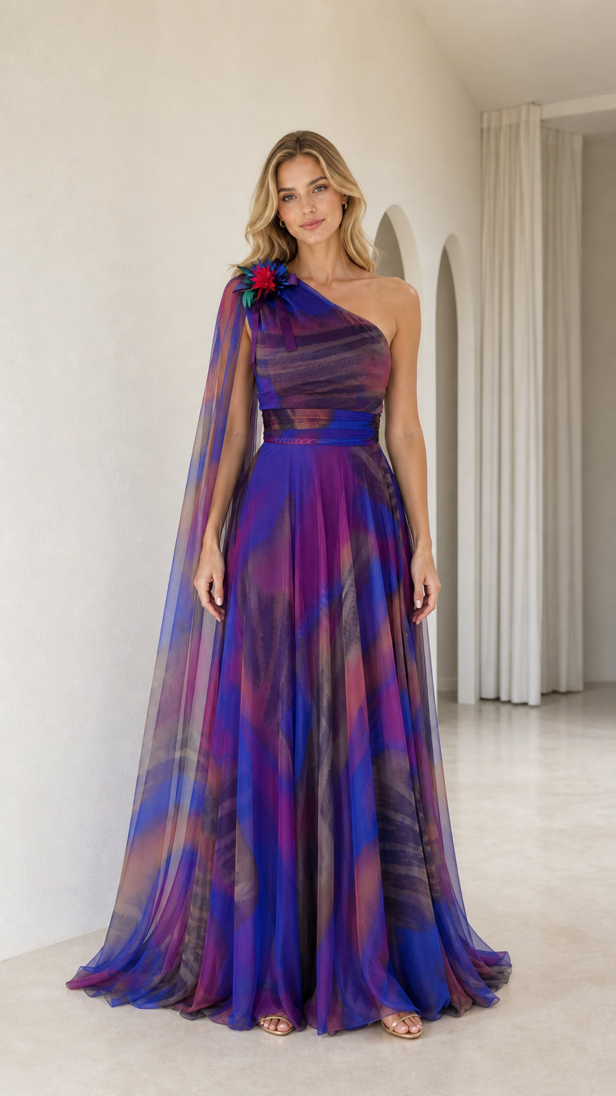 Front view of royal blue multi one-shoulder maxi evening gown with long sheer cape and floral corsage