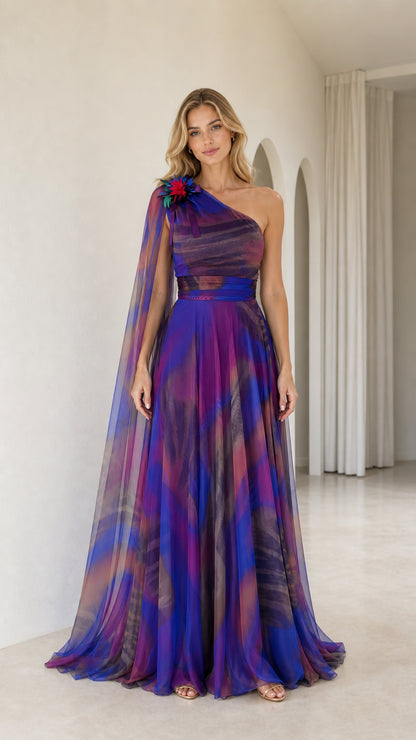 Front view of royal blue multi one-shoulder maxi evening gown with long sheer cape and floral corsage