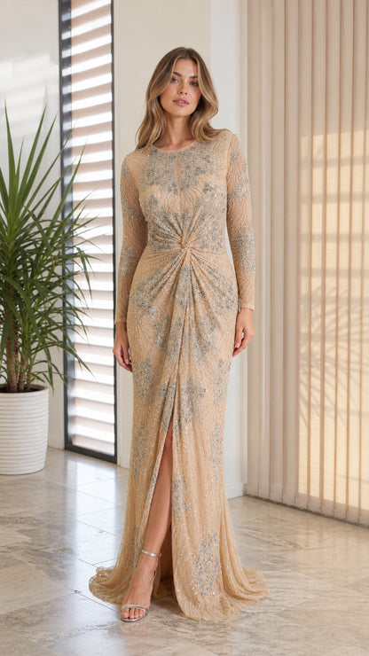 Front full-length view of model wearing champagne embellished long-sleeve gown with twist waist and slit